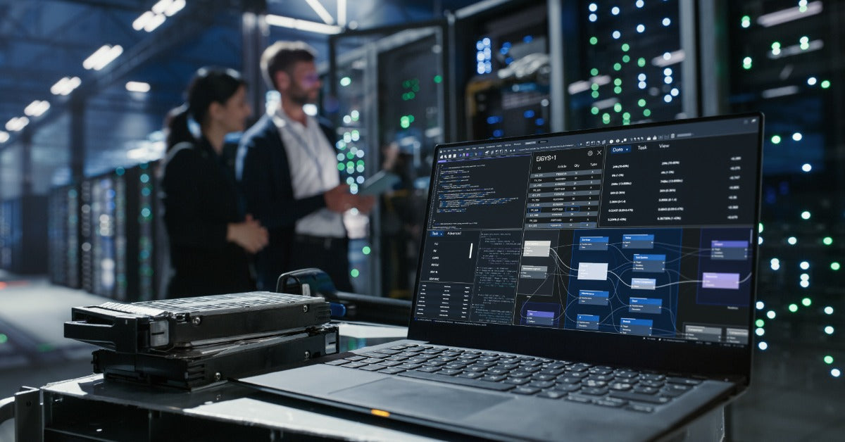 A laptop and stacked hard drives on a cart in a server room, with two people talking near equipment.