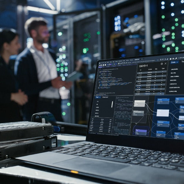 A laptop and stacked hard drives on a cart in a server room, with two people talking near equipment.
