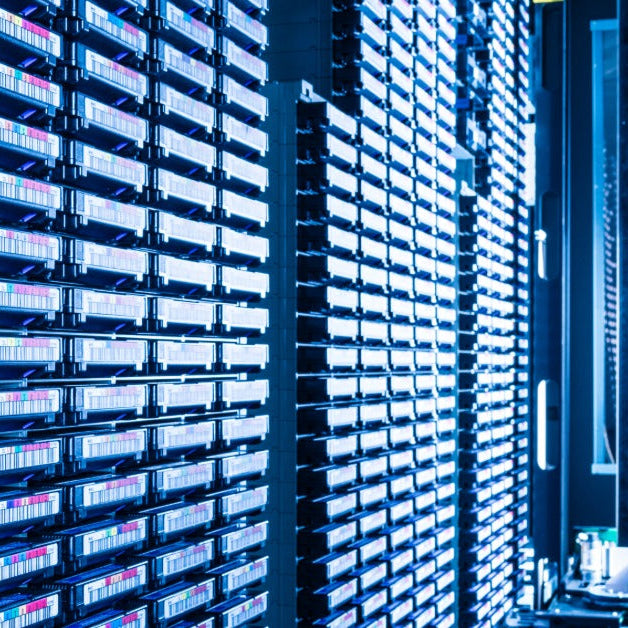 Rows of magnetic tape cartridges lining tall storage racks inside a blue-lit data storage room with narrow aisles.